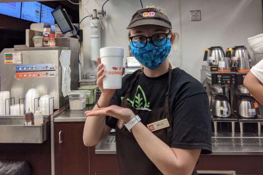 Working at the Dunkin Donuts drive thru in St. Clair, senior Bella Woodford poses with a coffee. Woodford wears protective gear like gloves and a mask due the coronavirus outbreak. We have to sanitize our lockers where we keep our stuff and we have a plastic guard covering half the window. If we feel any symptoms, we have to call HR and we need to stay out for two weeks, Woodford said.